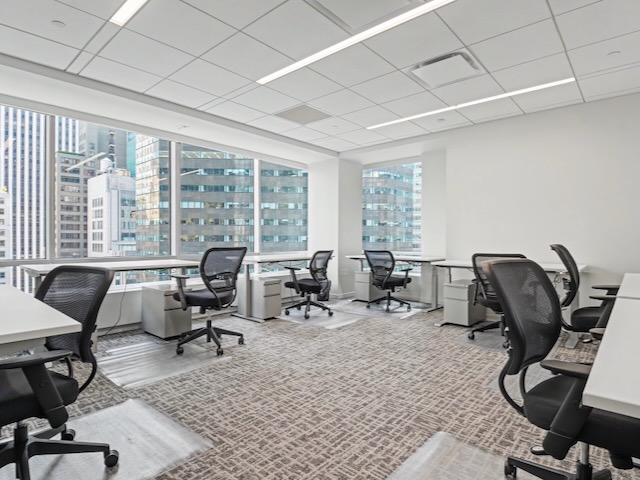 Modern office with empty desks and chairs, large windows, and a city view with tall buildings outside.
