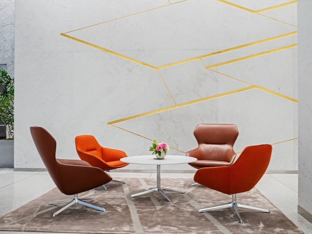 Four modern chairs surround a round table with a flower vase on a rug, set against a marble wall with gold lines.