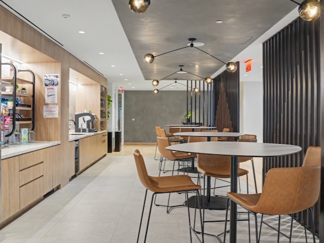 Modern office break room with high tables, brown chairs, snack area, and geometric ceiling lights.