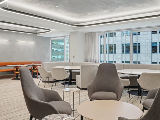 Modern office lounge with gray chairs, round tables, and large windows showing city buildings.