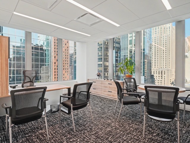 Modern office with black chairs, wood desks, large windows, and a city skyline view in the background.