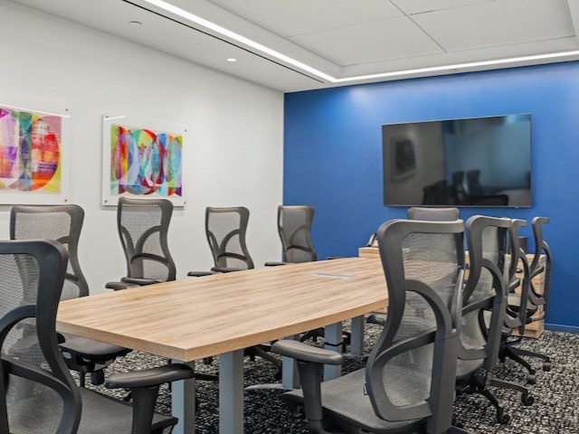 Modern conference room with a wooden table, mesh chairs, blue accent wall, TV, and abstract art.