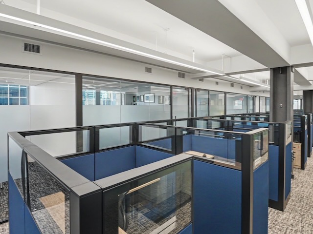 Modern office with blue cubicles, glass walls, and overhead lighting in an open workspace setting.