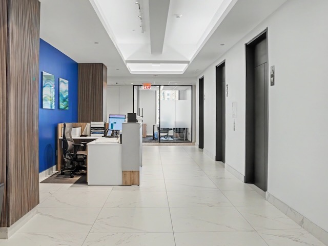 Modern office hallway with white floors, blue accent wall, elevators, and desks with computers on the left.