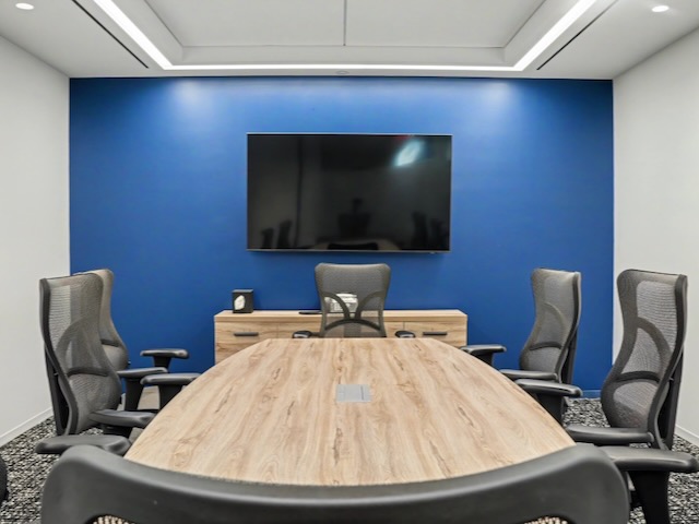 Modern conference room with a wooden table, black chairs, blue accent wall, and a large screen.