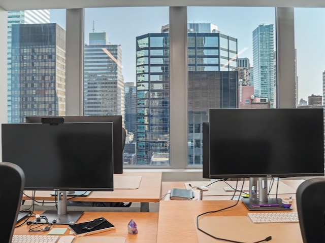 Empty modern office with computer monitors and large windows showing city skyscrapers outside.