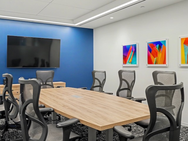 Modern conference room with a wooden table, eight chairs, wall art, and a flat-screen TV on a blue wall.