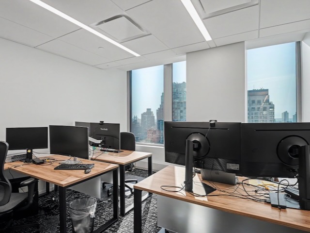 Modern office with wooden desks, computer monitors, chairs, and large windows overlooking city buildings.