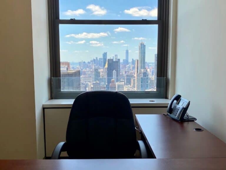 Empty office desk and chair by a window overlooking a city skyline with tall buildings and blue sky.