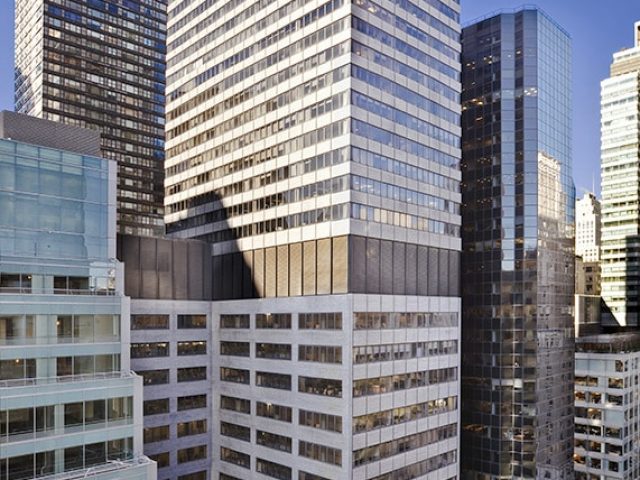 A tall office building with reflective glass windows stands among other high-rise structures on a clear day in a downtown area. An intersection is visible in the lower right corner.