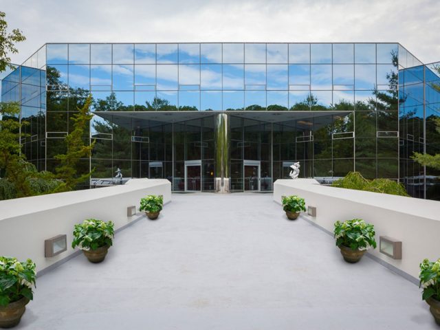 Modern glass building with mirrored windows, viewed from a walkway lined with potted plants.
