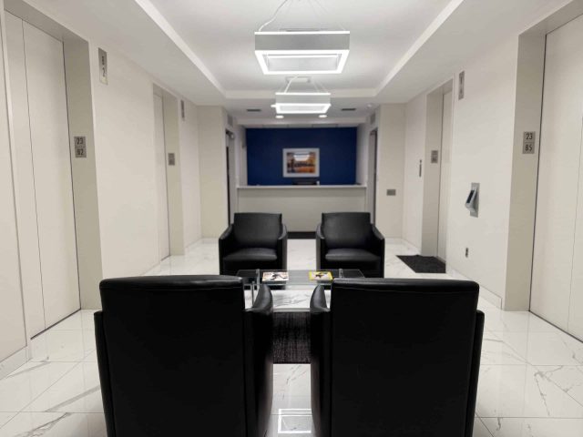 Modern elevator lobby with four black chairs arranged around a small table, white walls, blue accent wall, and elevators on both sides.