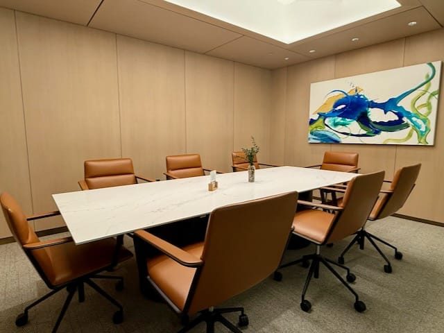 A modern conference room with a white marble table, six brown leather chairs, light wood walls, and an abstract painting. A vase with greenery sits on the table.